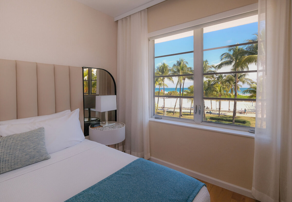 A guest room with a bed, nightstand, and window showing a beach view with palm trees.