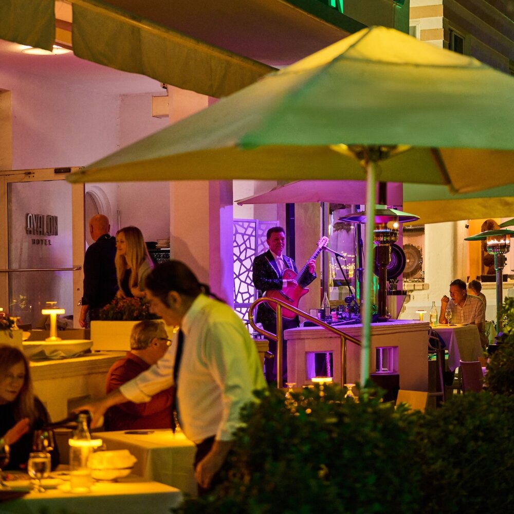 A nighttime view of a hotel bar with outdoor seating, a musician playing guitar, and patrons dining.