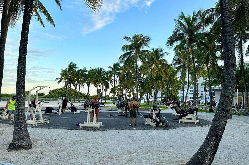 A beachside outdoor gym with exercise equipment and people working out, surrounded by palm trees and a blue sky.