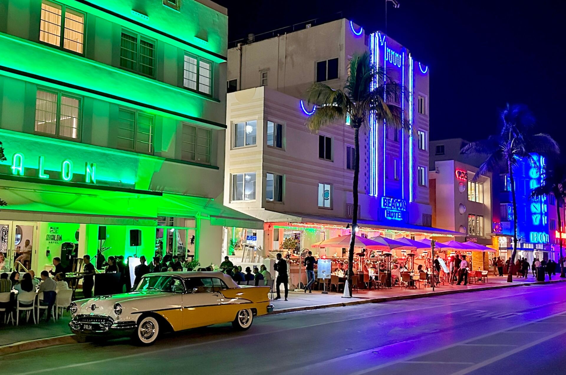 A nighttime view of a hotel exterior with green and blue neon signs, palm trees, and a vintage car parked in front of a restaurant patio.