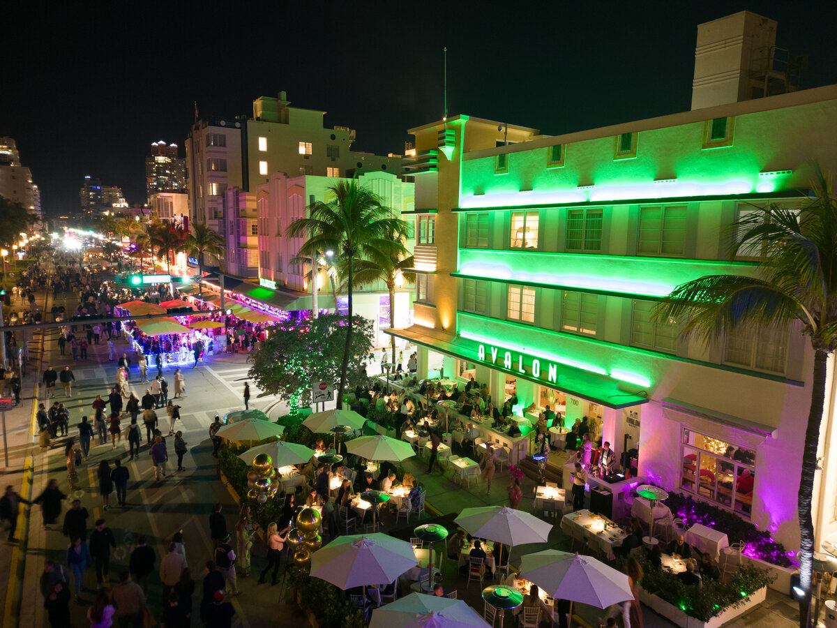 A nighttime view of a building with green and purple lights, surrounded by people and palm trees.