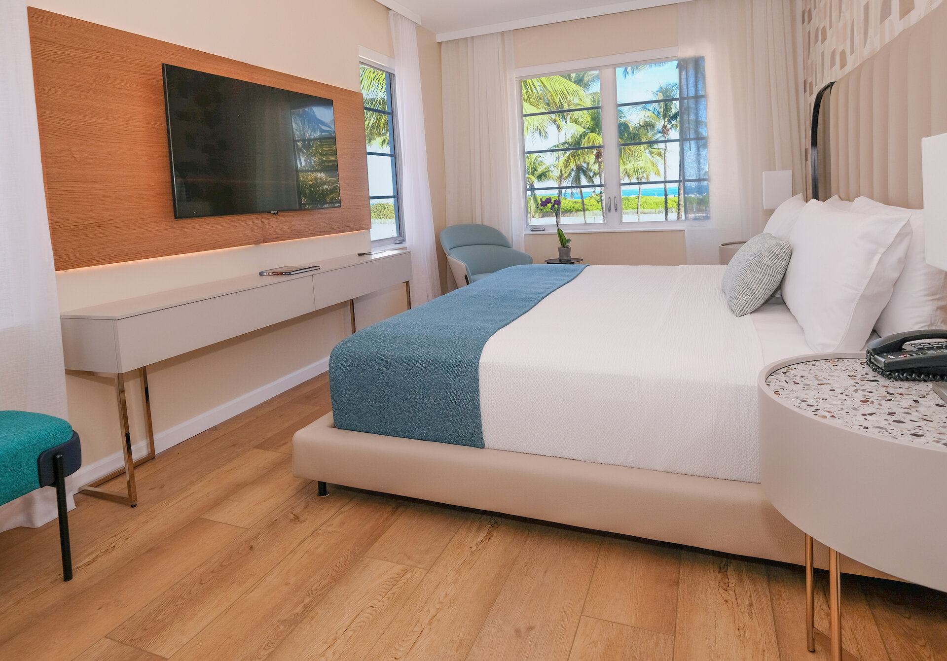 A hotel room with a large bed, TV, desk, and chair, with a view of palm trees outside the window.
